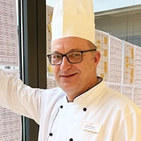 Armin Storm, Küchenchef in Kochjacke und Mütze, vor Menüplänen am Fenster.
