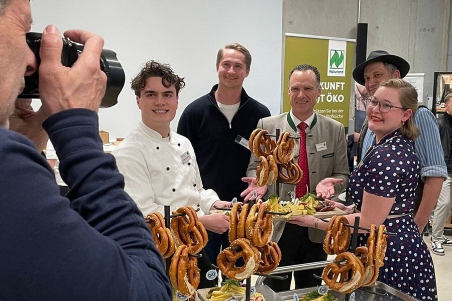 Mehrere Personen stehen zusammen und halten eine Platte mit Lebensmitteln. Im Vordergrund fotografiert jemand die Gruppe.